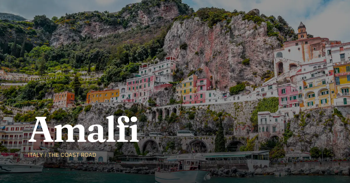 A daytime photo of the Amalfi cliffside town, pastel buildings stacked up a steep rocky cliff above the boats and water, with the place name Amalfi set large in a serif over the bottom band and an Italy kicker - the strong-photo move for travel.